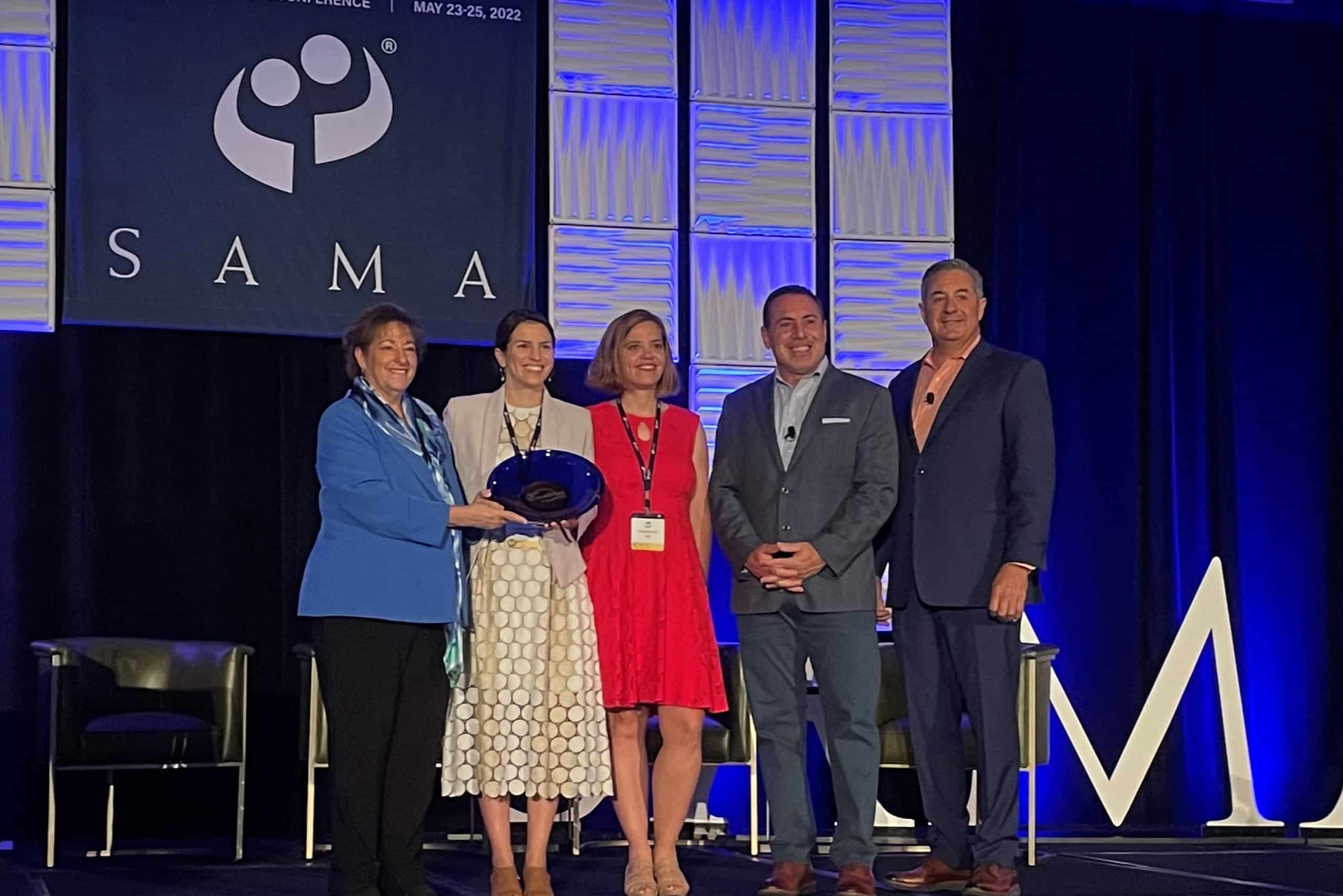 Five people on stage, smiling. Three hold awards in business attire. SAMA backdrop and event details in blue lighting behind them.