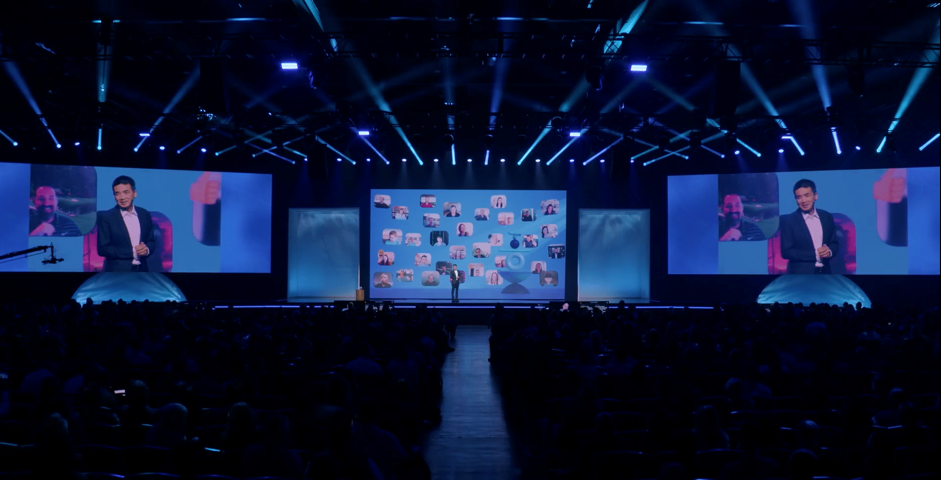 A presenter on stage with blue lighting; screens display his image and peoples photos. Auditorium filled with attendees.