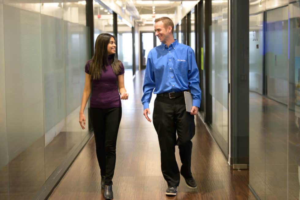A woman in a purple top and man in blue shirt walk and talk in a glass-walled office hallway with wooden floors and ceiling lights.