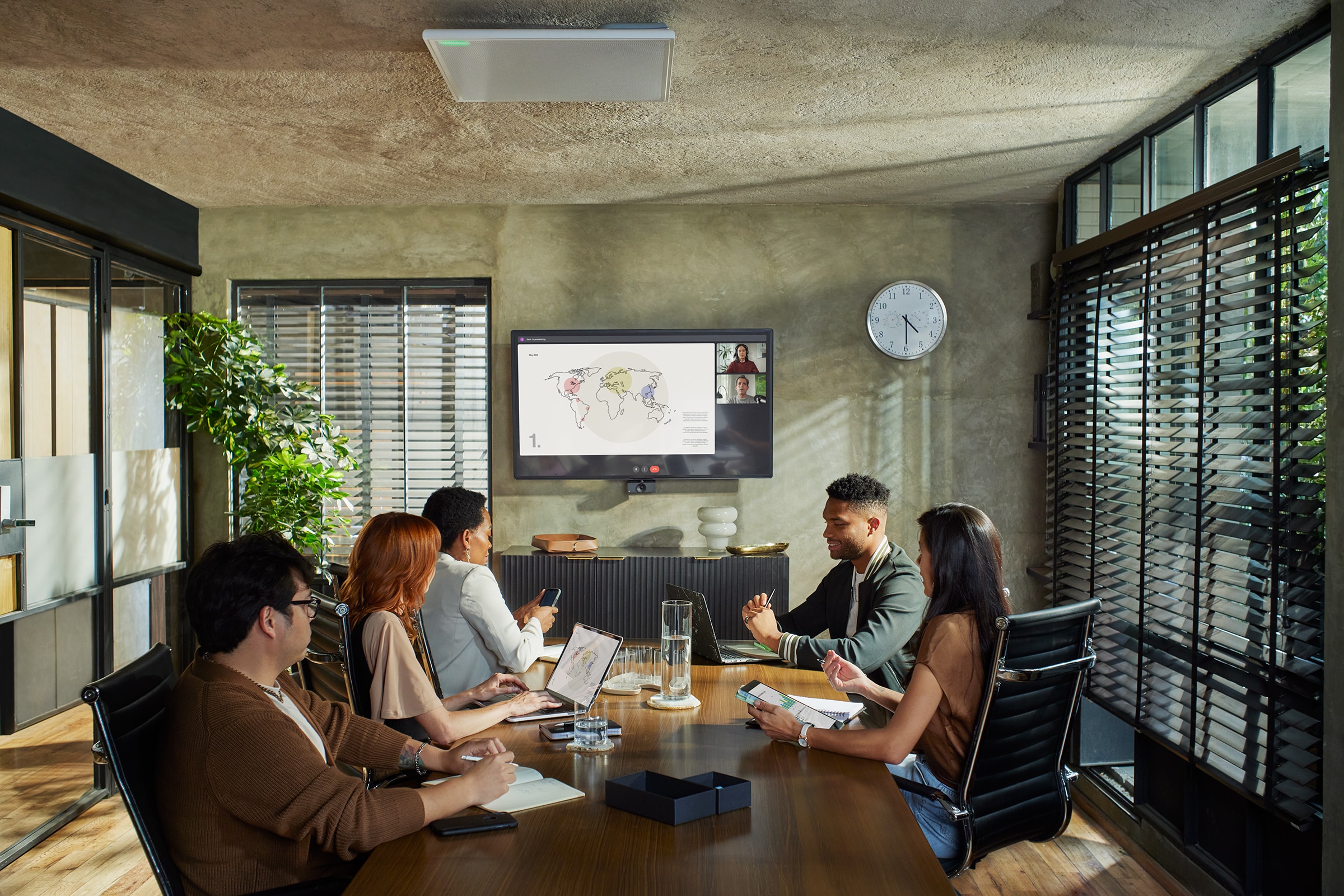 Six diverse individuals sit at a conference table, engaging in a video call. Sunlight filters through windows, highlighting room plants.