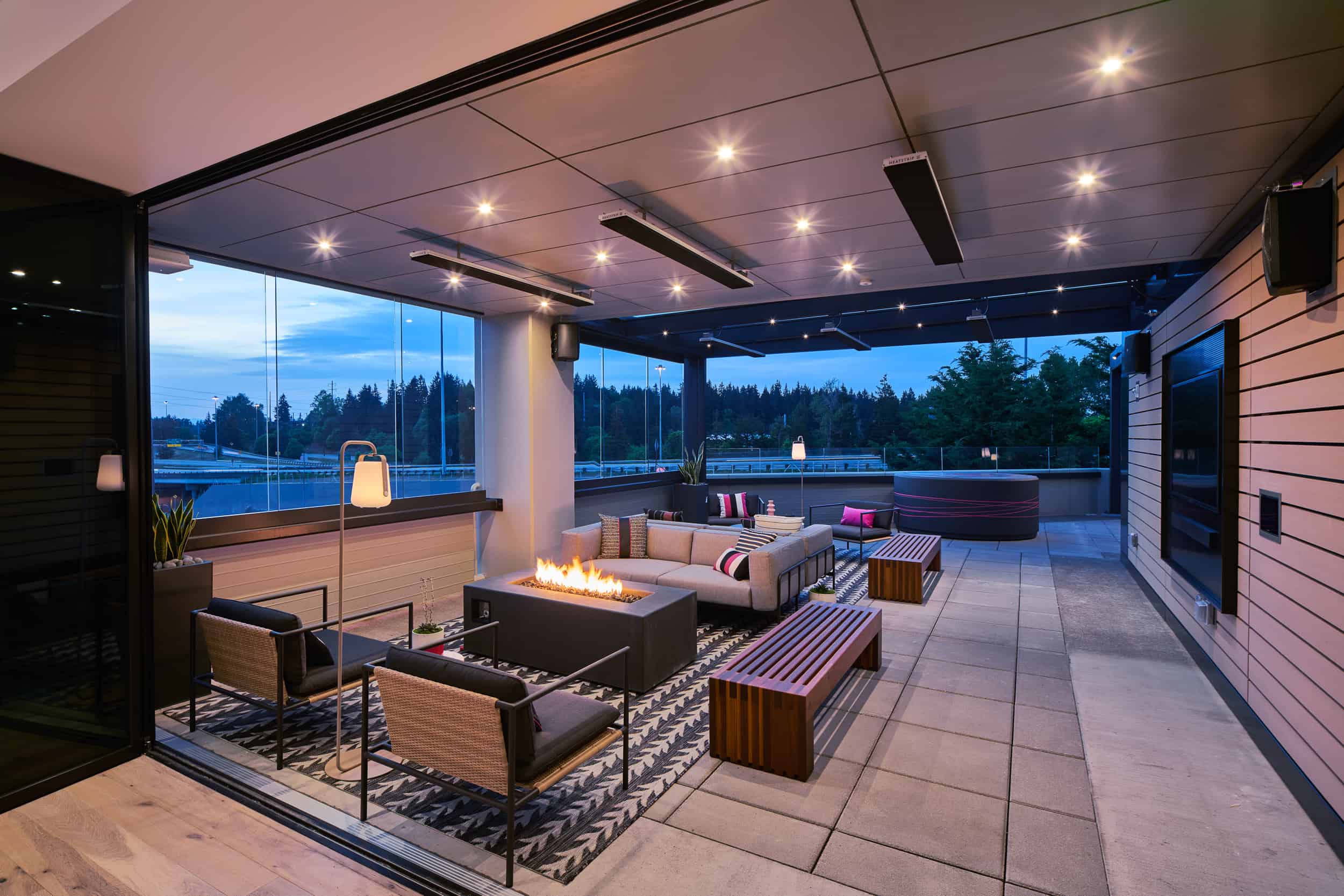 Modern patio with cozy seating around a fire pit on a rug; lights overhead, scenic water view in the background.