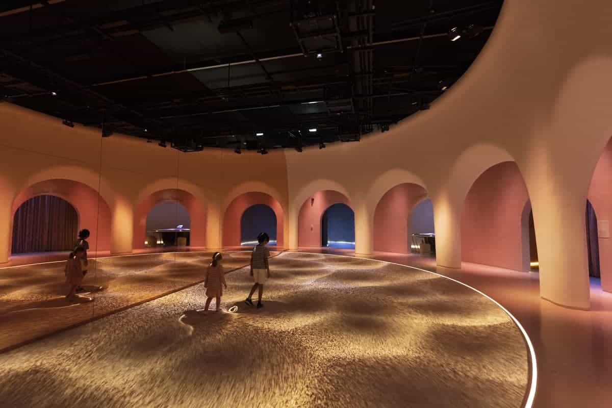 A modern, spacious room with high ceilings. Illuminated floor patterns shift as three people walk across, adding scale and movement.