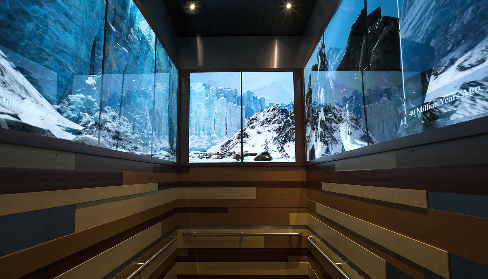 Elevator with screens showing snowy mountains across panels; walls have striped geometric patterns and side handrails.