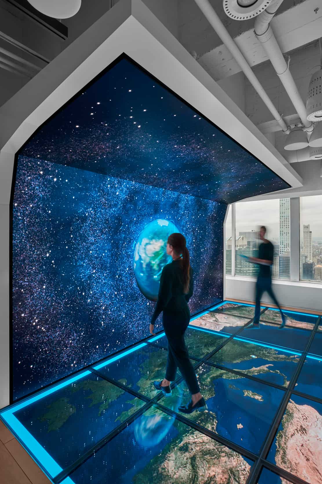 Woman approaches screen showing starry space with Earth; man blurred nearby. Floor glows with maps, ceiling pipes above.