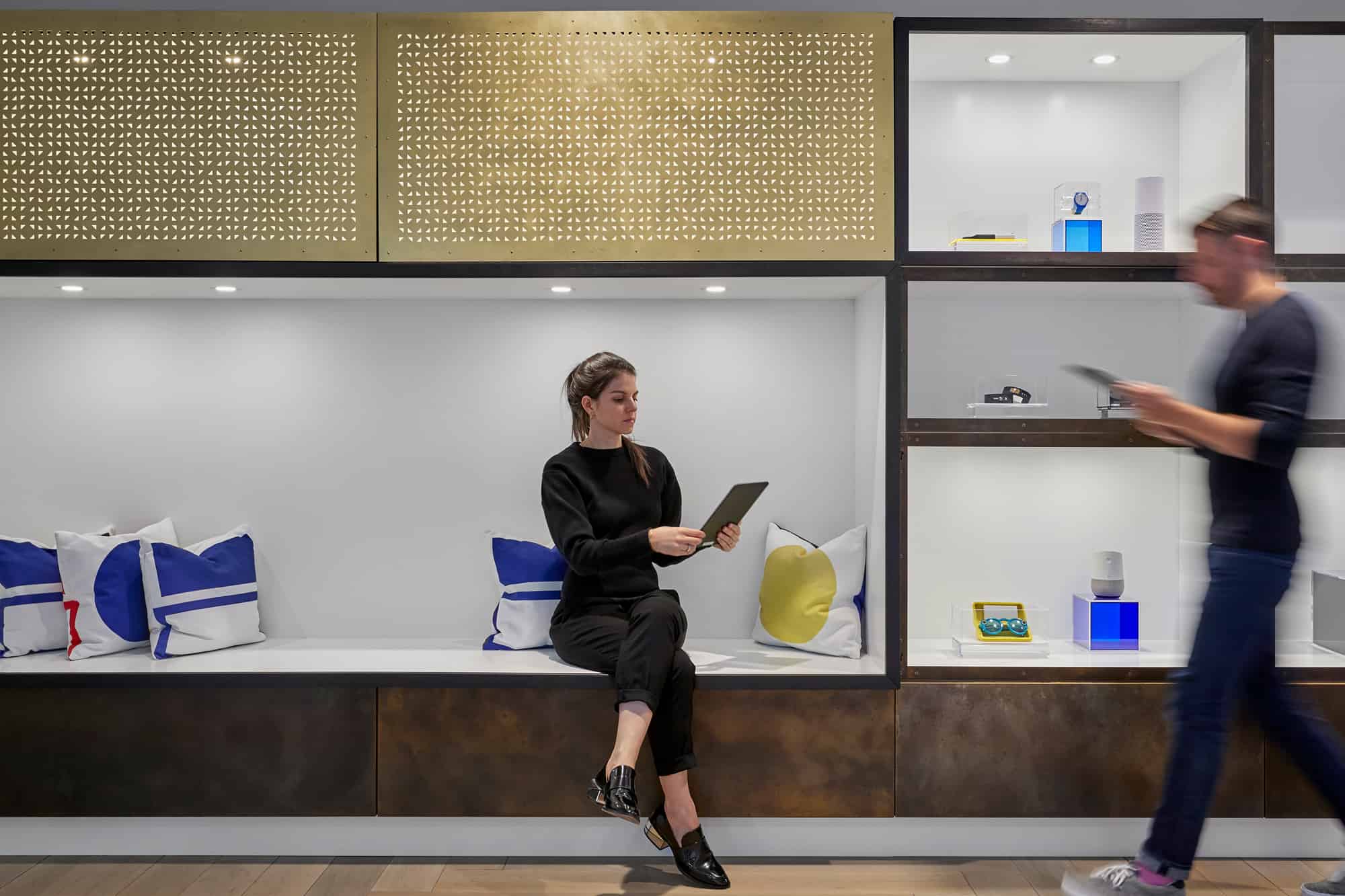Woman on bench reads tablet in modern space; shelves with decor around. Man walks by with tablet, motion blur adds dynamic calm.