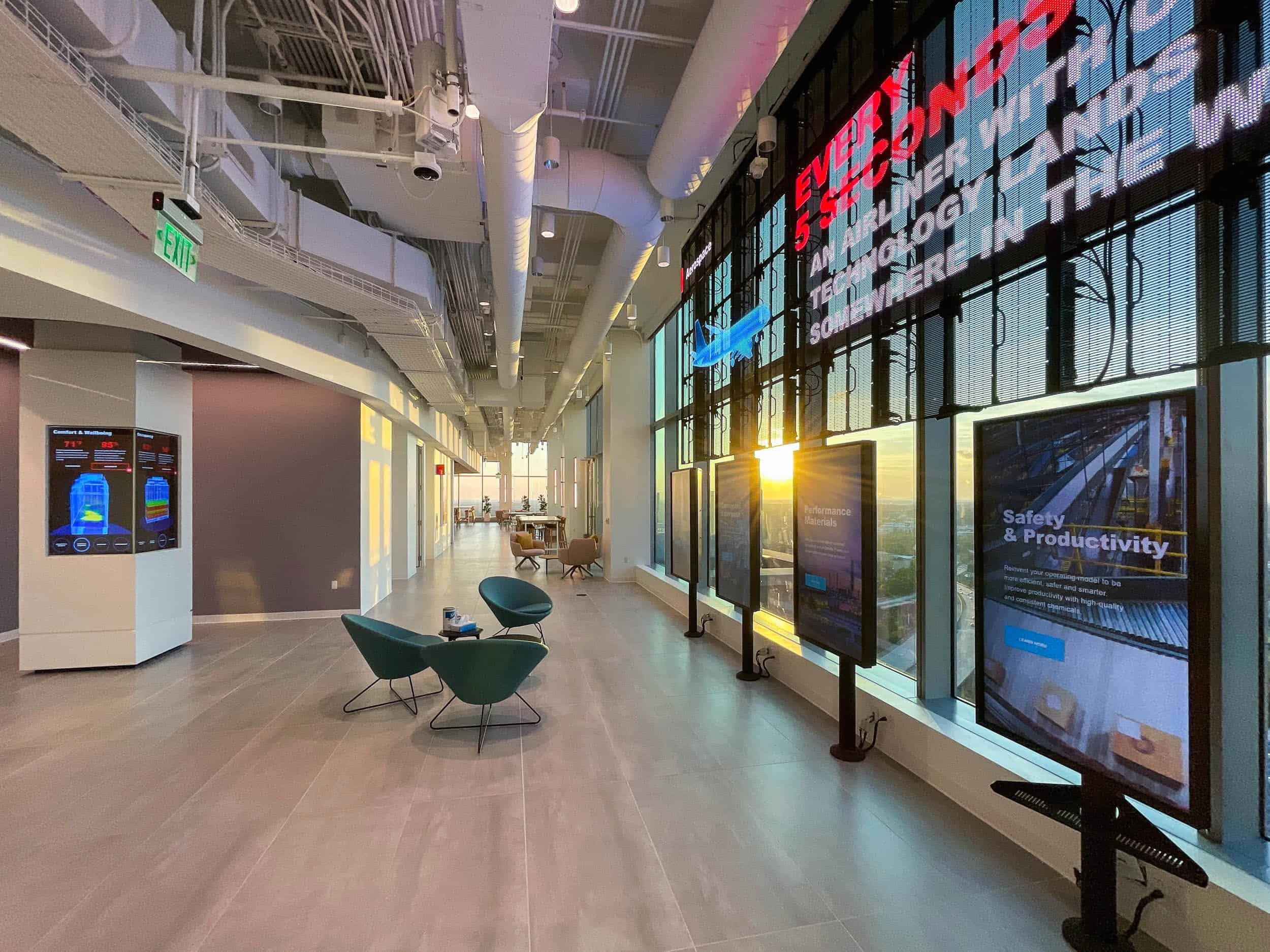 Modern lobby: digital displays with tech content, teal chairs & a small table by entrance, long hallway, bright ceiling lights.