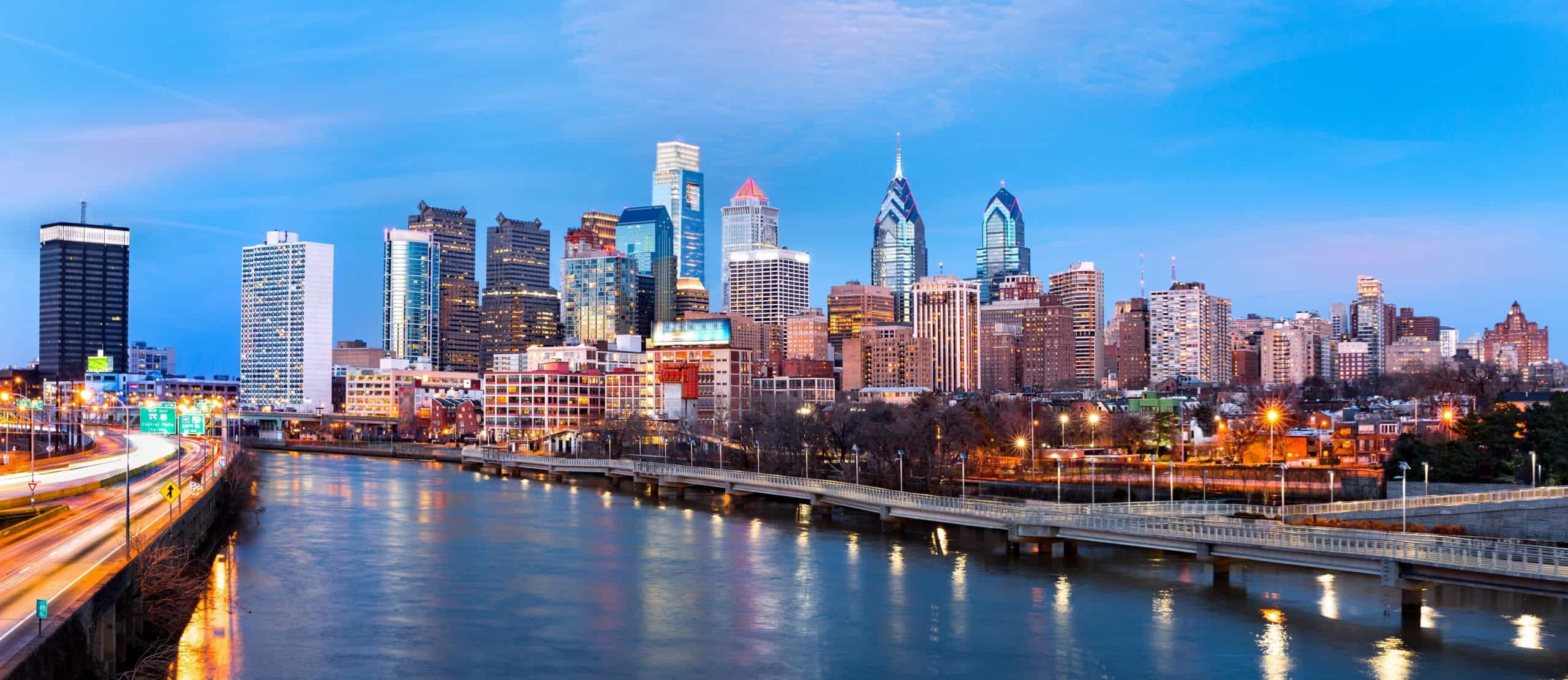 Philadelphia skyline at dusk with skyscrapers illuminated, Schuylkill River in foreground, sky blue and pink; lights reflecting on water.