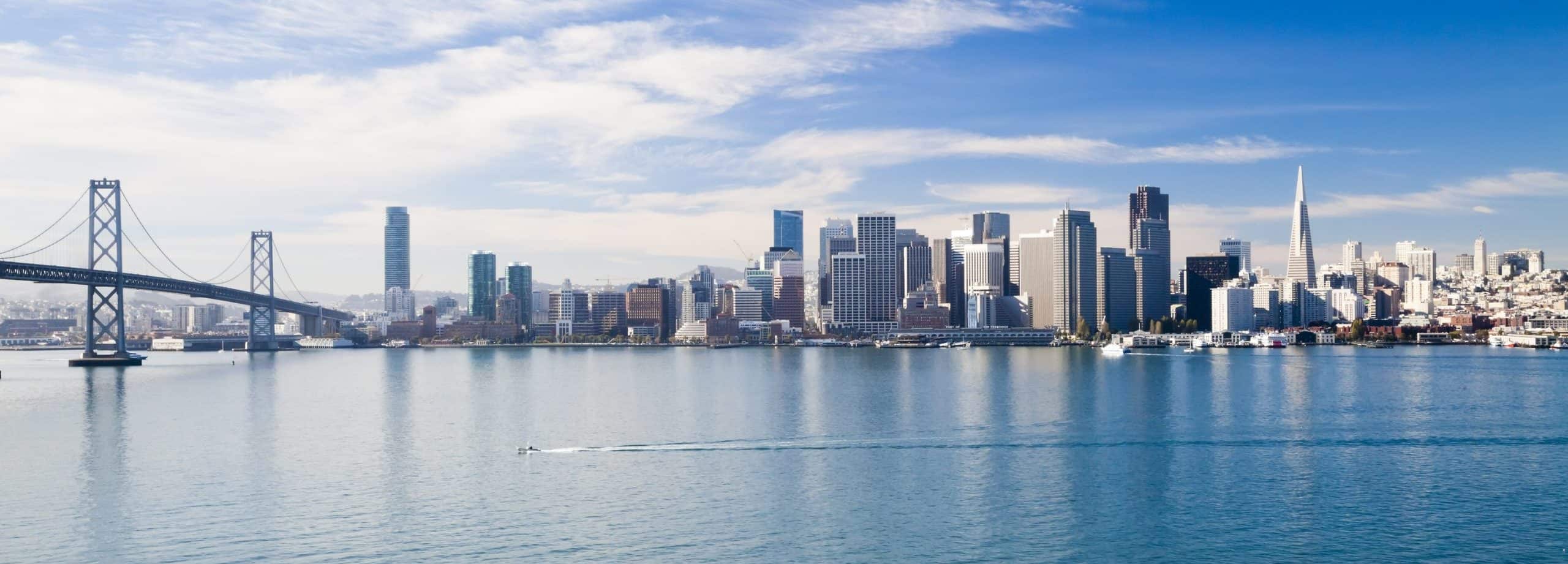 San Francisco skyline with Bay Bridge, mirrored on calm waters. Boat ripples under blue sky with scattered clouds.