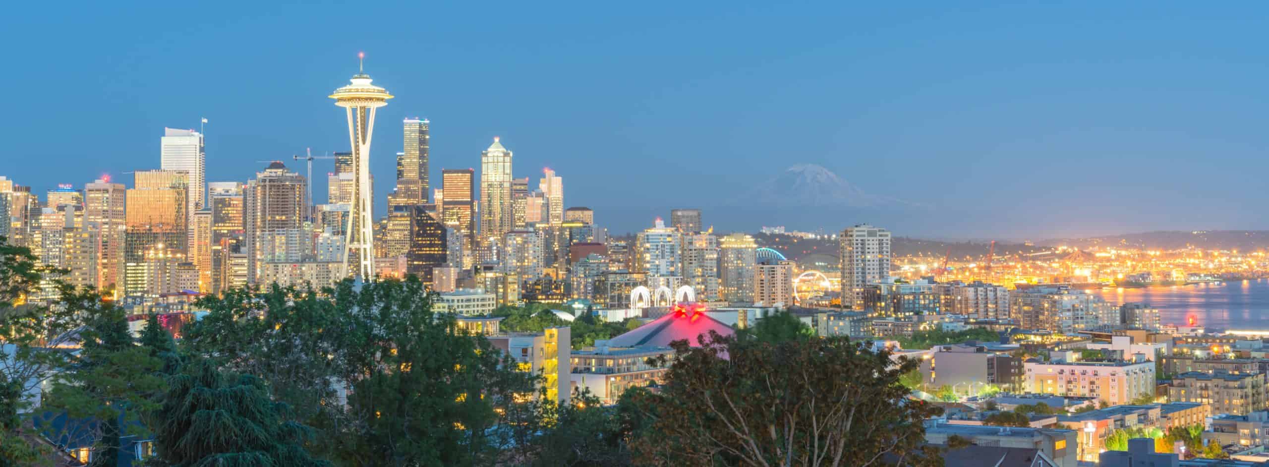 Seattle skyline at dusk: Space Needle shines, city aglow, Mount Rainier faintly visible. Foreground trees frame the scene.