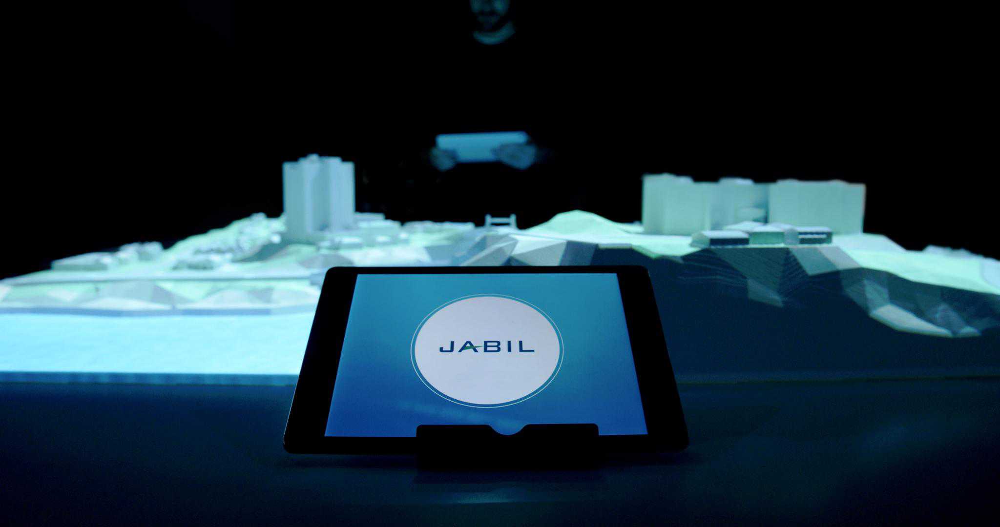 Tablet shows JABIL in foreground; blurred 3D city model and a person holding a tablet in the dark background.