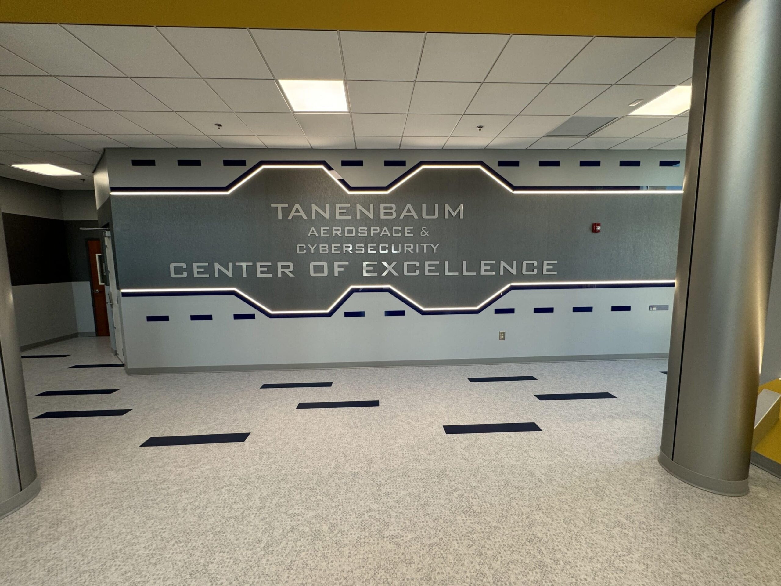 Place the Tanenbaum Aerospace & Cybersecurity Center of Excellence sign in a modern hallway, carpeted with navy and recessed lighting.
