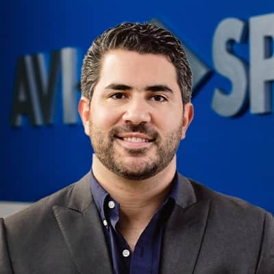 Man with short dark hair and a beard smiling, wearing a dark blazer over a collared shirt. Blue background with partial text.