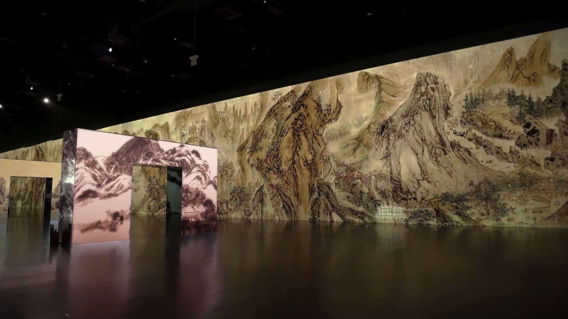 A dimly lit room showcases landscape paintings on freestanding panels, featuring mountains and trees for an immersive experience.