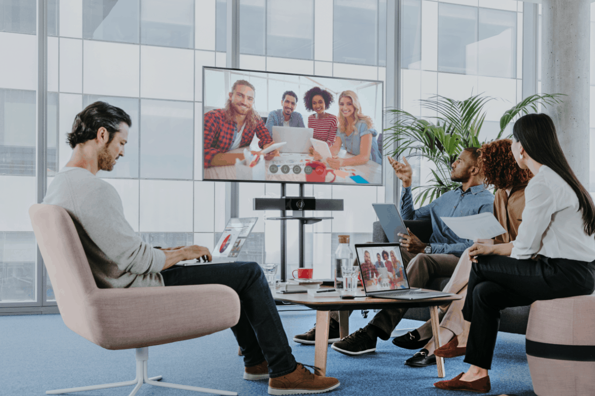 Office team on video call: group around table with laptops/documents, screen shows 4 smiling; one with laptop in foreground.