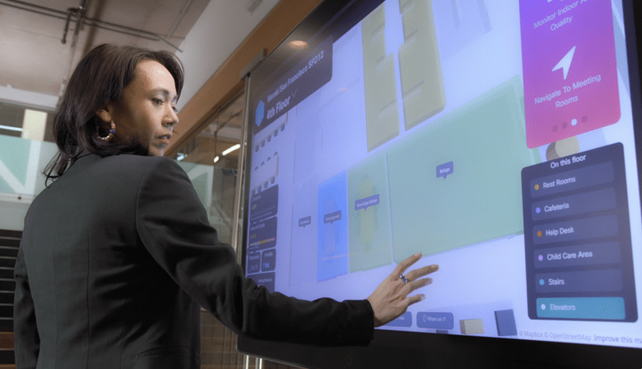 A person in a black blazer uses a large touchscreen to navigate a digital building map in a modern office setting.