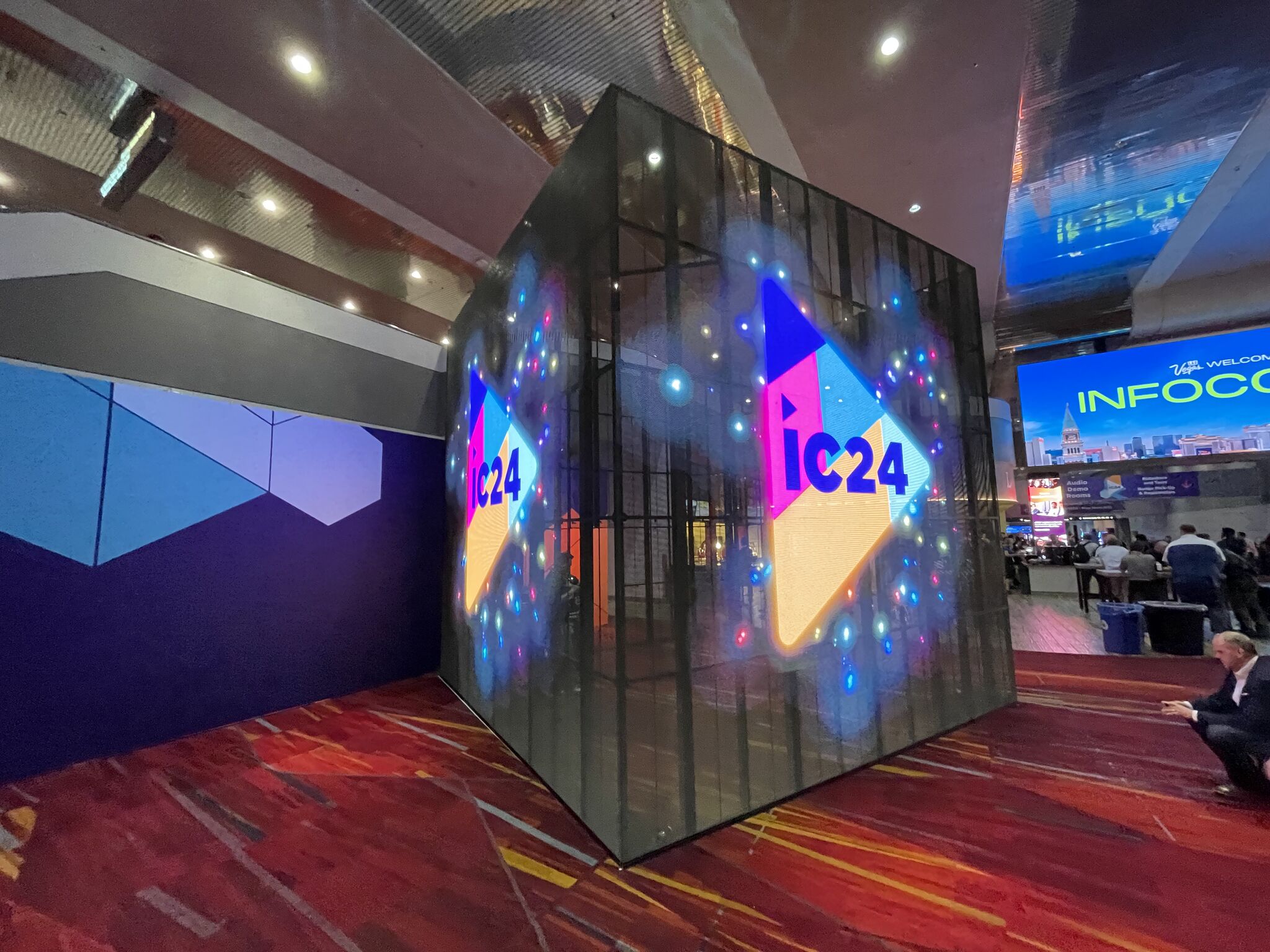 A cubic display with IC24 in colorful lights fills the hall, surrounded by people and booths. Reflective ceiling amplifies vibrancy.