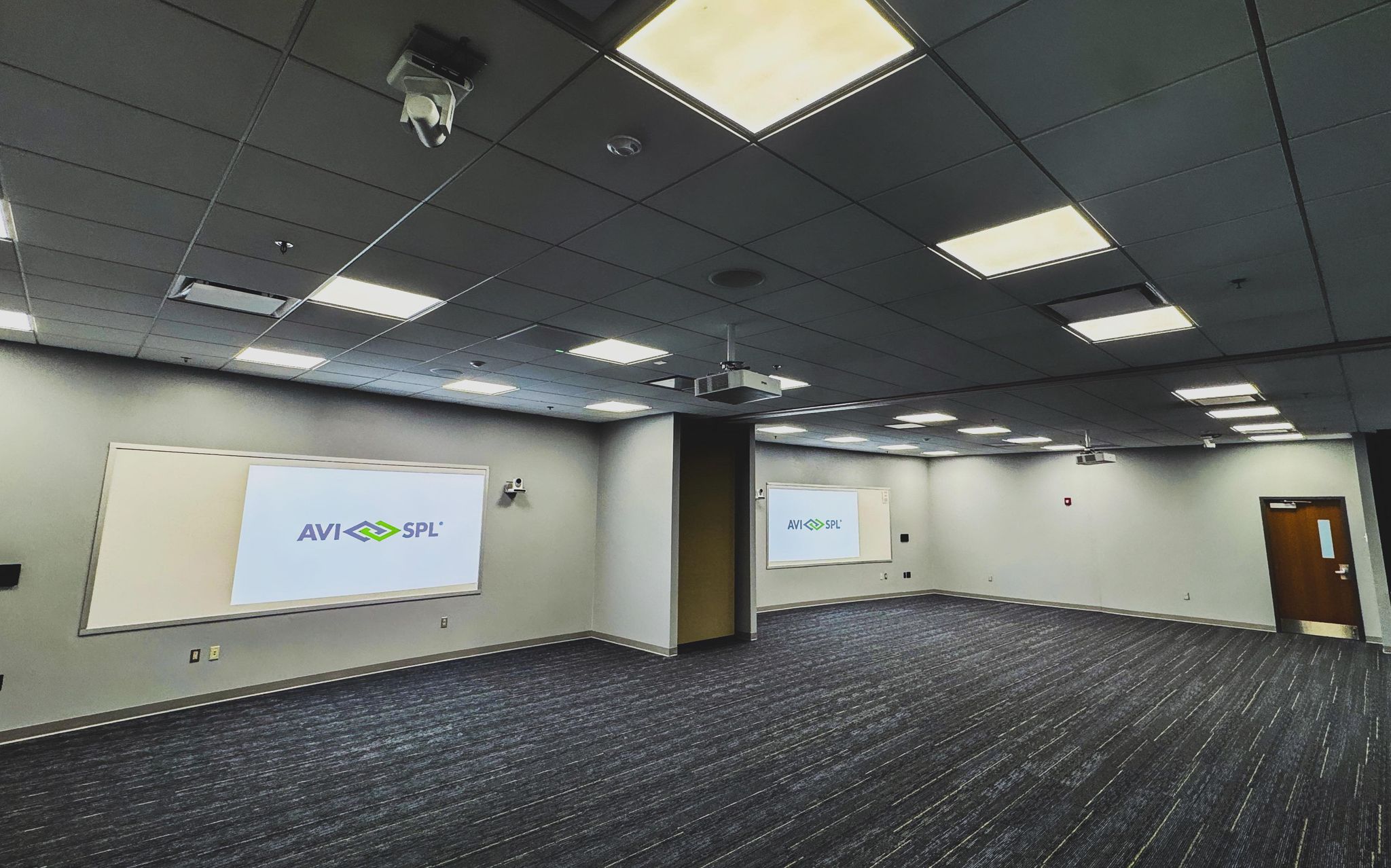 Modern conference room with two screens showing AVI-SPL logo, carpeted floor, recessed ceiling lighting, wooden door on right.