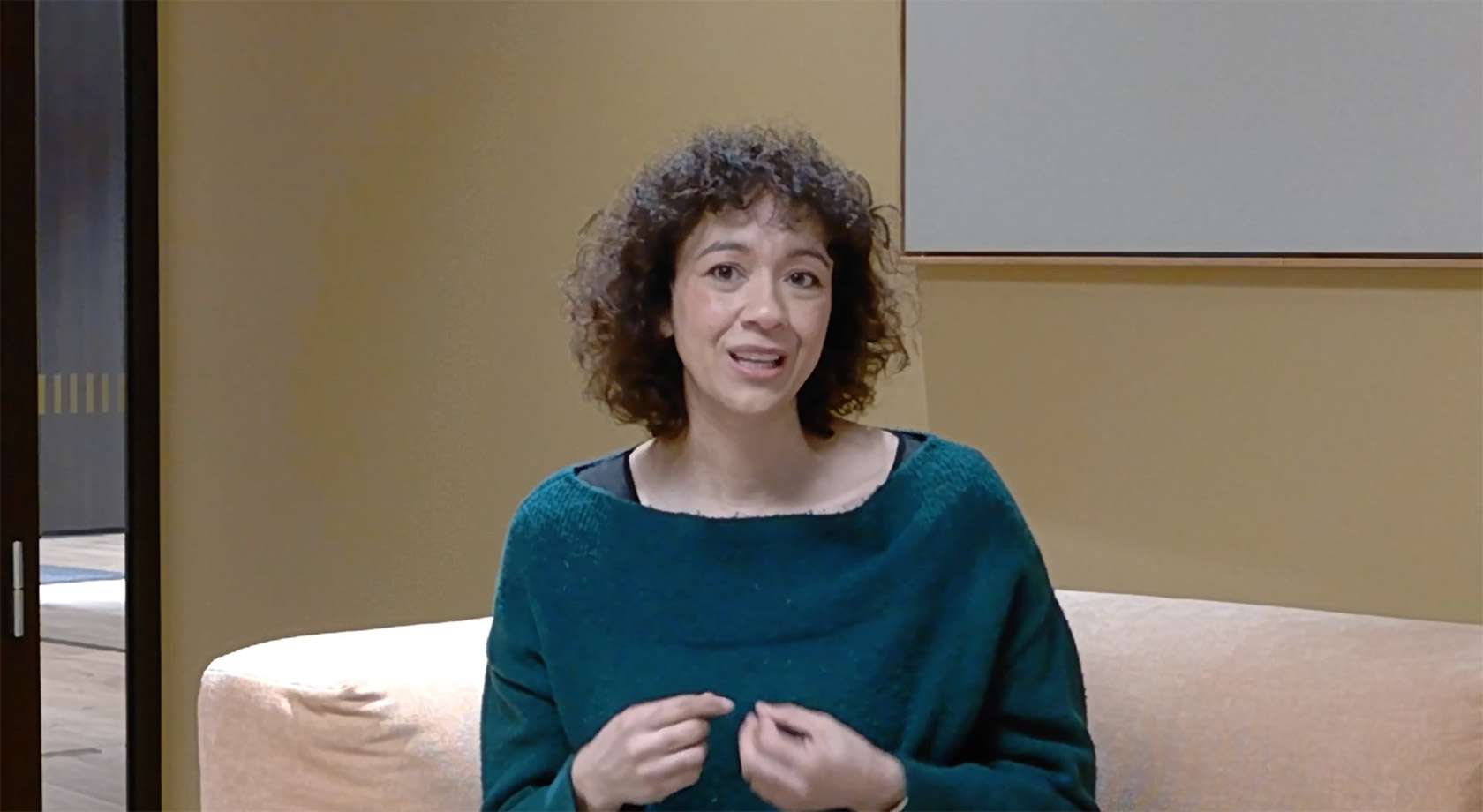 A curly haired woman sat on a meeting room sofa, talking to the camera.