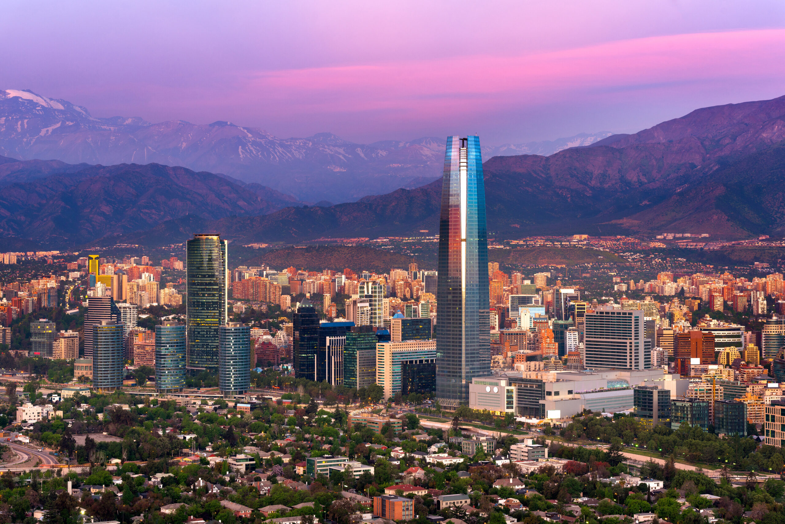 Panoramic view of Santiago at sunset: skyscrapers, lush greenery, Andes mountains under a pink-purple sky.