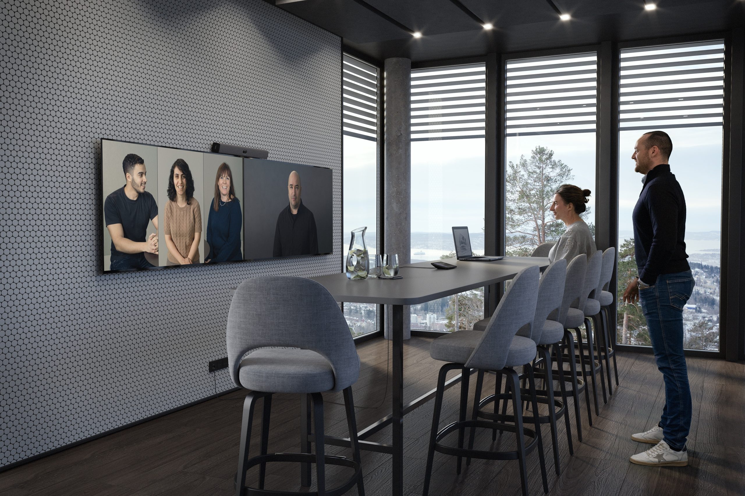 Two in a modern conference room on a video call; screen shows four. Water pitcher, glasses on table. Large windows, scenic view.