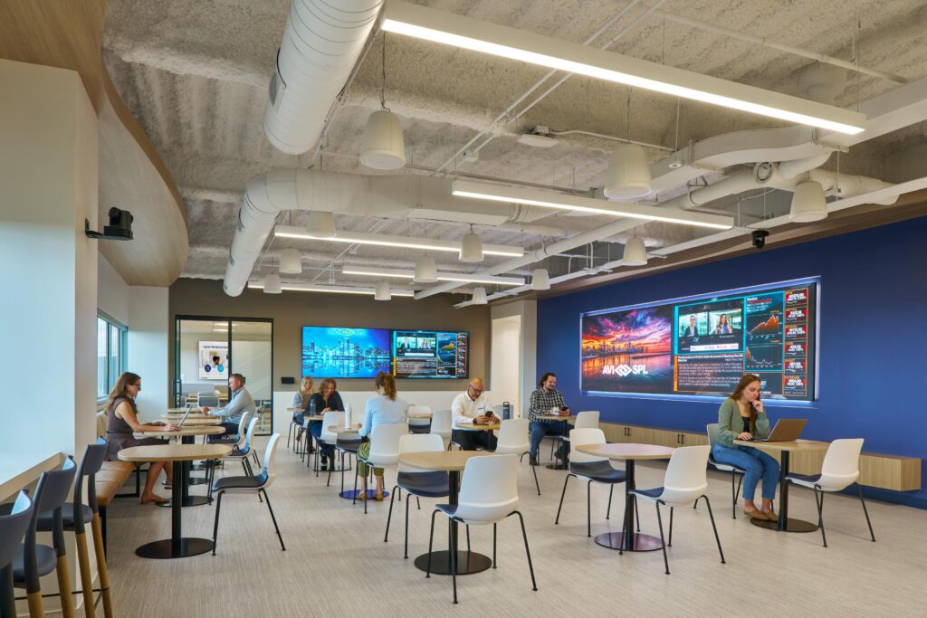 Modern office lounge: people working on laptops, discussing. Digital screens on walls, white furniture, exposed ducts.